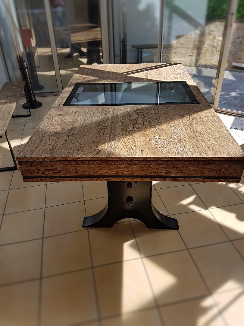 La beauté noire du pied tulipe en métal de cette table-billard ATELIER trône sur le carrelage grès de cette pièce aux nombreuses baies vitrées. La noblesse de la finition chêne brossé huilé ressort grace à la partie centrale vitrée du plateau qui montre le tapis de jeu de billard couleur gris ardoise.